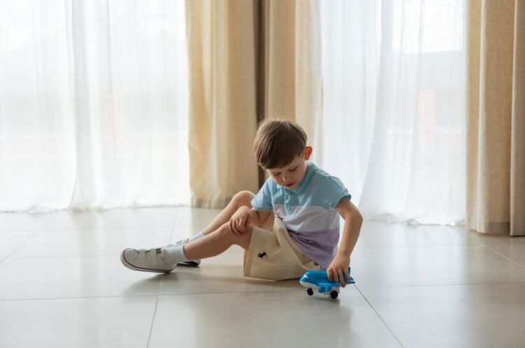 A young boy sits on the floor playing with a blue toy airplane in a sunny room.