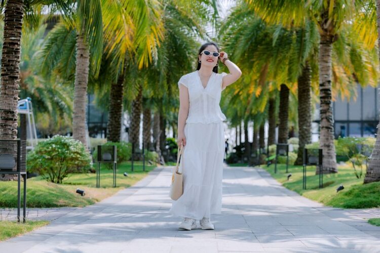 Stylish woman in white dress and sunglasses walking down a sunny palm-lined pathway.
